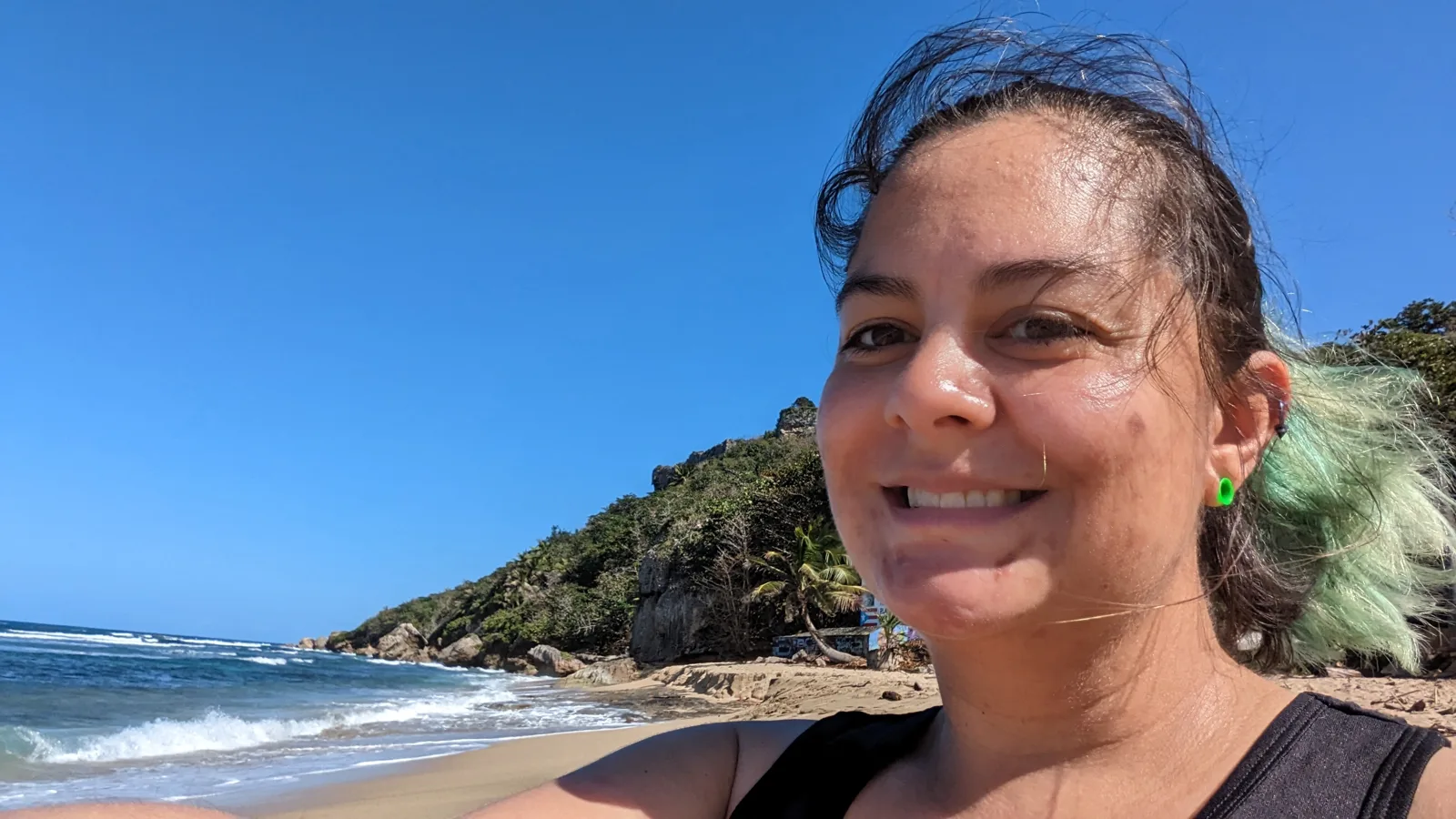 Abigail smiling in a selfie on a sunny beach with a green headland behind