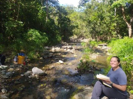 Abigail sitting beside a cobble-bed stream, writing in a field notebook