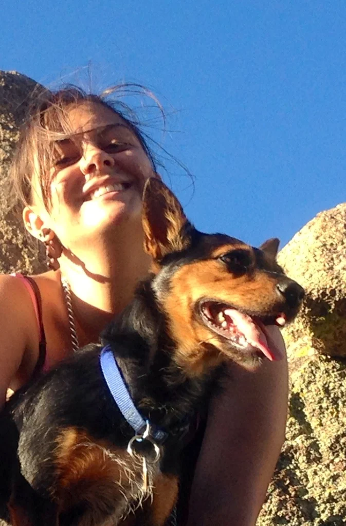 Abigail smiling in the sun with Marsala, nicknamed Wolverine, her small black-and-brown Lancashire heeler, against a rocky outcrop